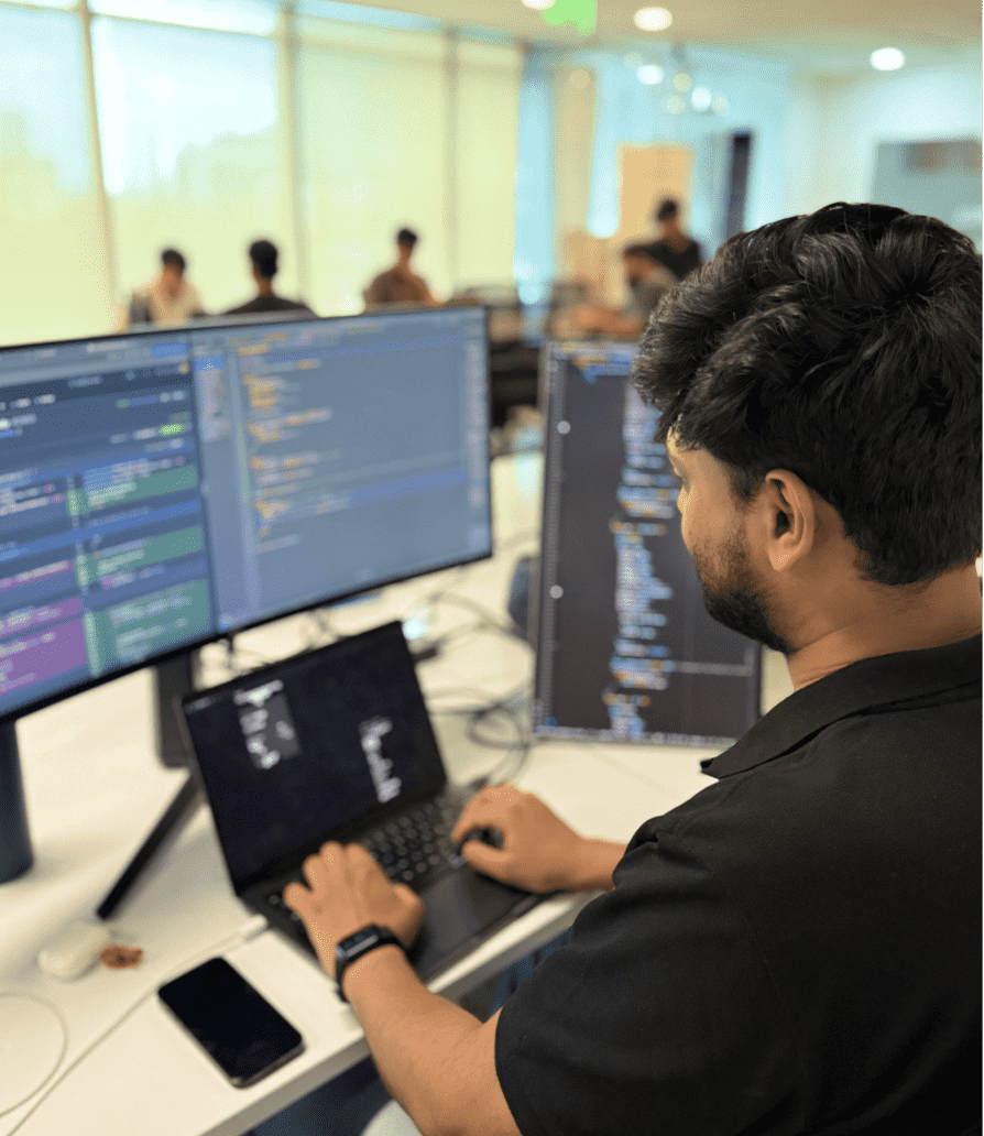 Engineer at a TestMu AI office writing test code across a laptop and two monitors