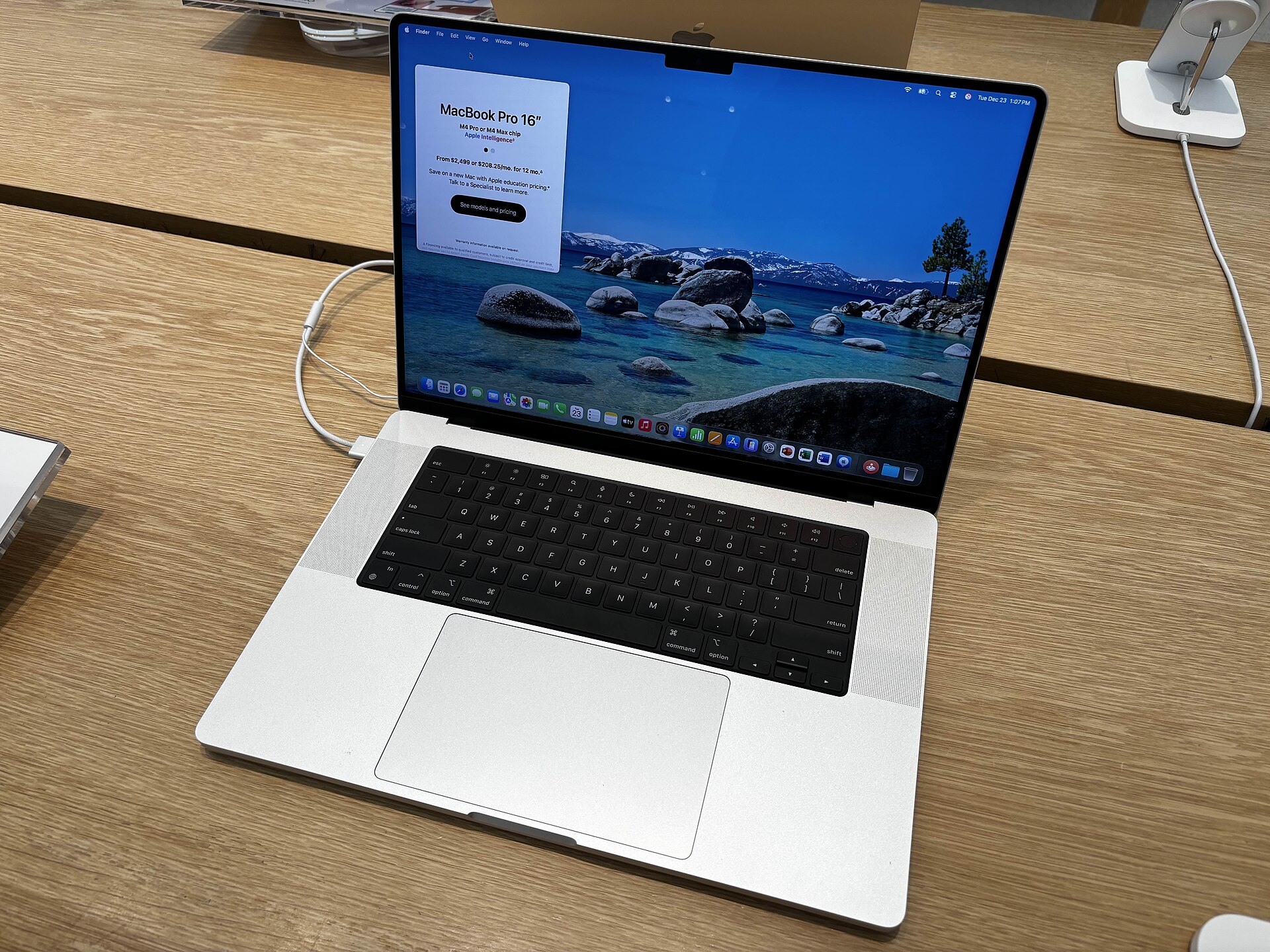 An open silver 16-inch MacBook Pro on a wooden table showing a lake wallpaper